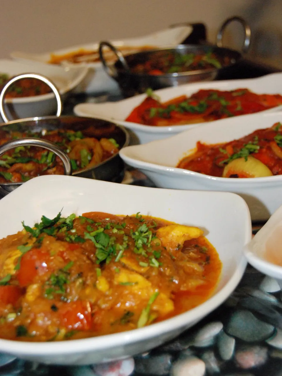 A row of our curries in white serving dishes and steel balti pans, sprinkled with fresh coriander, with a tomato and onion dish in the foreground.