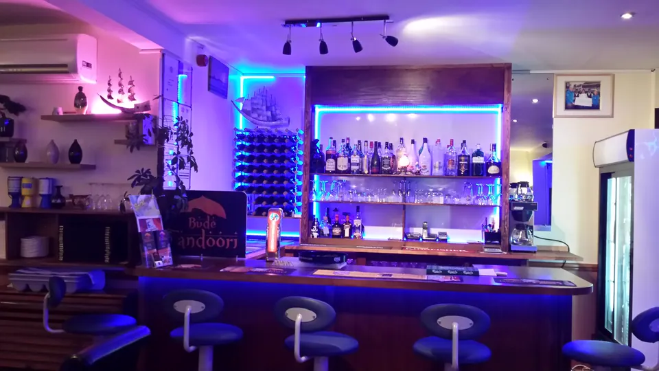 The bar: a wooden counter with stools, shelves of spirits and glasses lit in blue, and a wine rack beside it.
