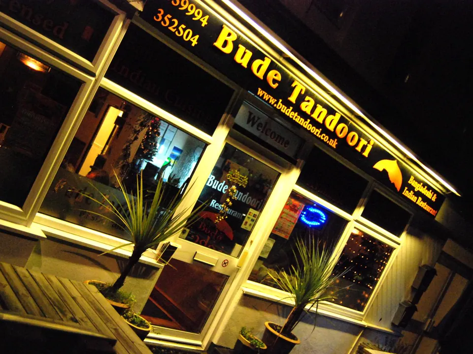 The Bude Tandoori shopfront on Burn View at night, with the lit sign above the door, two potted plants and a wooden bench outside.